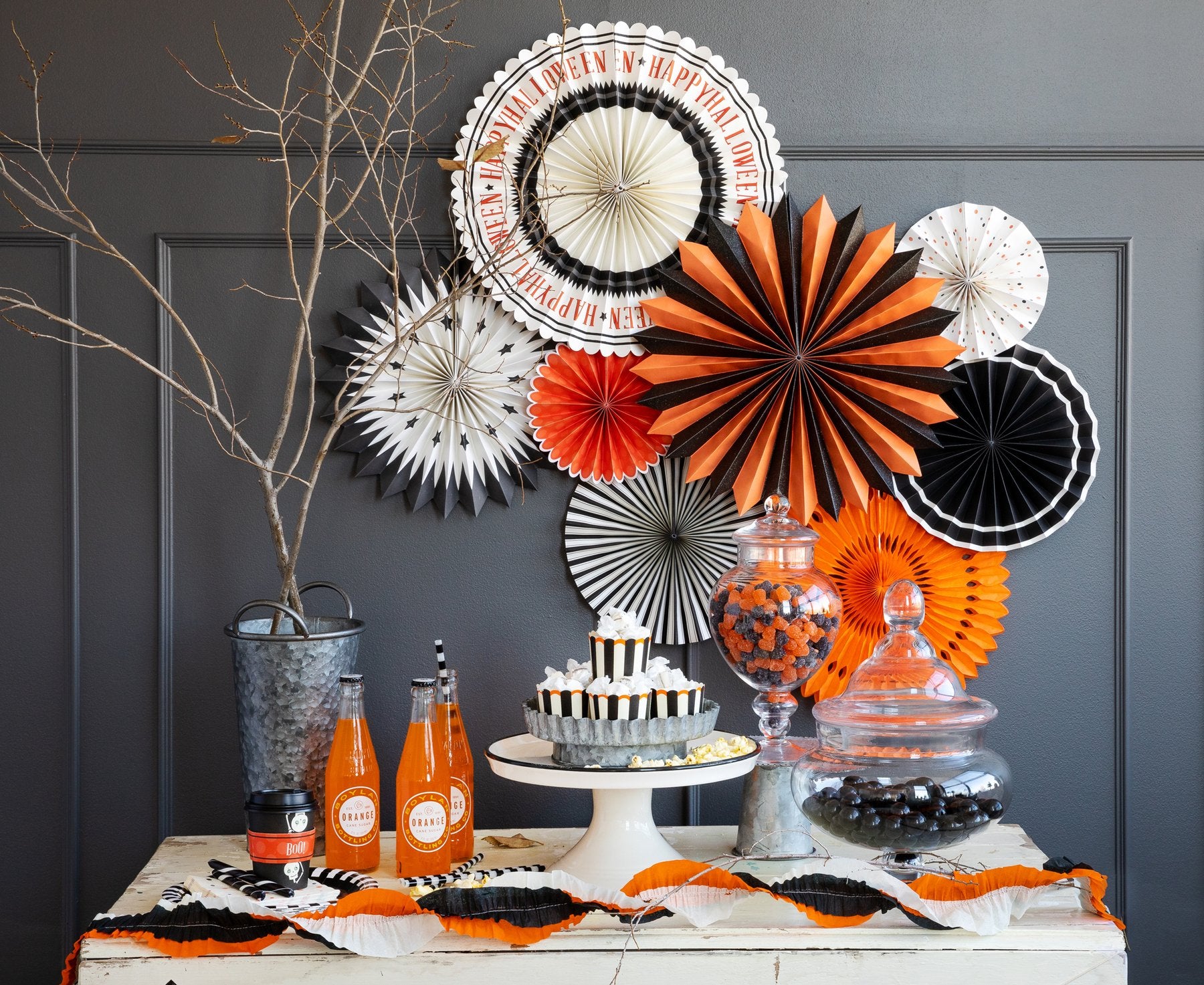 Halloween decor, different colored party fans, orange and black party fans, white party fans with black stars in front of dessert table.  Featuring black and white striped cupcakes, candy jars of gum-balls and orange soda with streamers