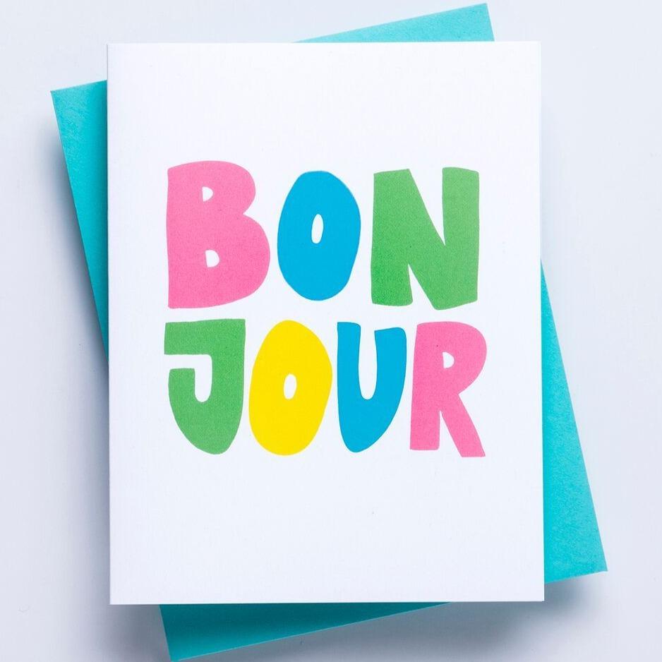 A white greeting card with the word 'Bonjour' displayed on the face in multicolored lettering with a blue enveloped