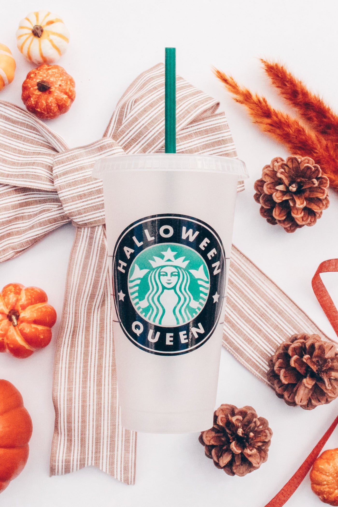 A frosted white reusable plastic Venti Starbucks cup with the Starbucks logo that reads Halloween Queen set among fall and Halloween type decor