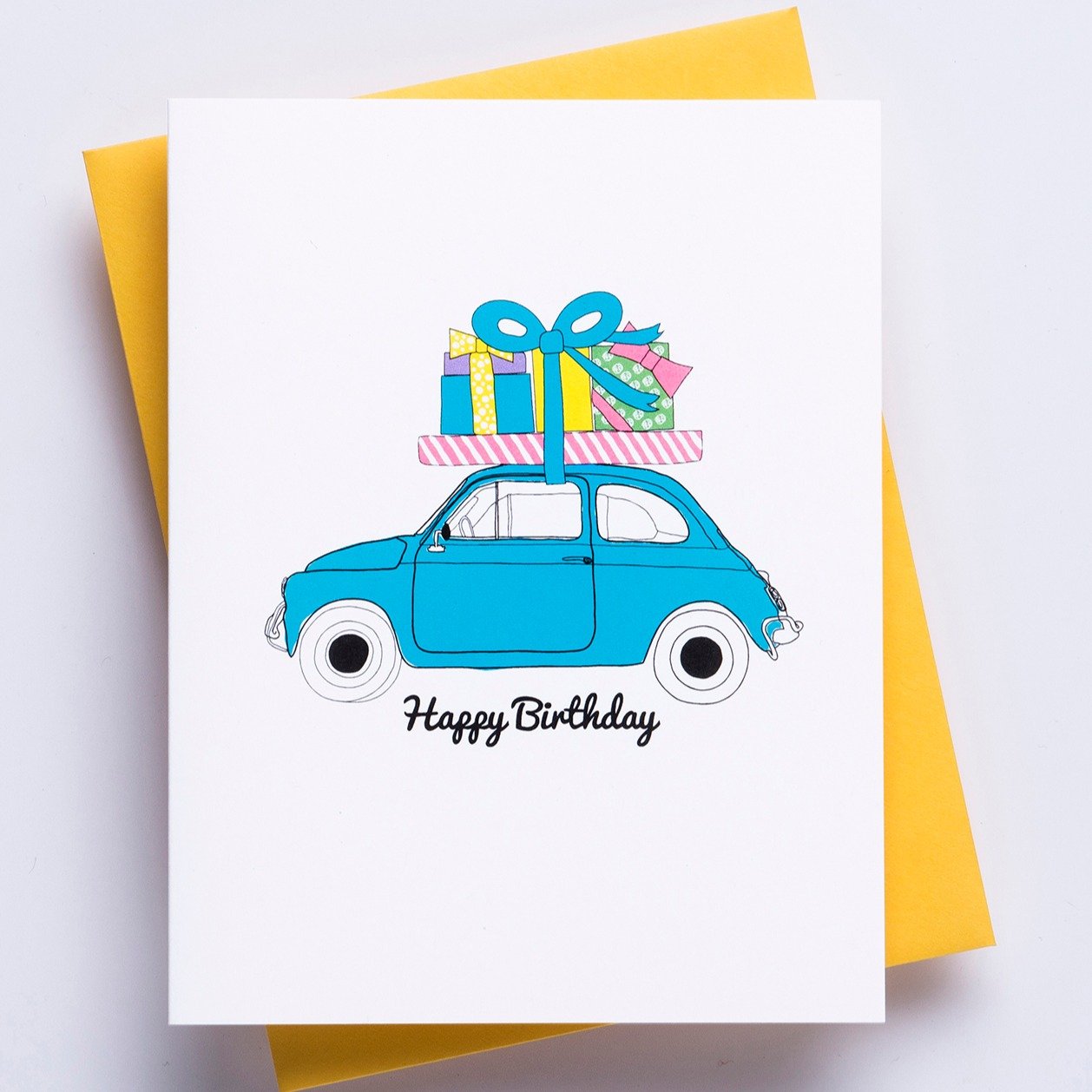 Happy Birthday Moto Greeting Card - A white greeting card with a image of a small car carrying birthday gifts on the roof and the words 'happy birthday' accompanied by a bright yellow envelope.