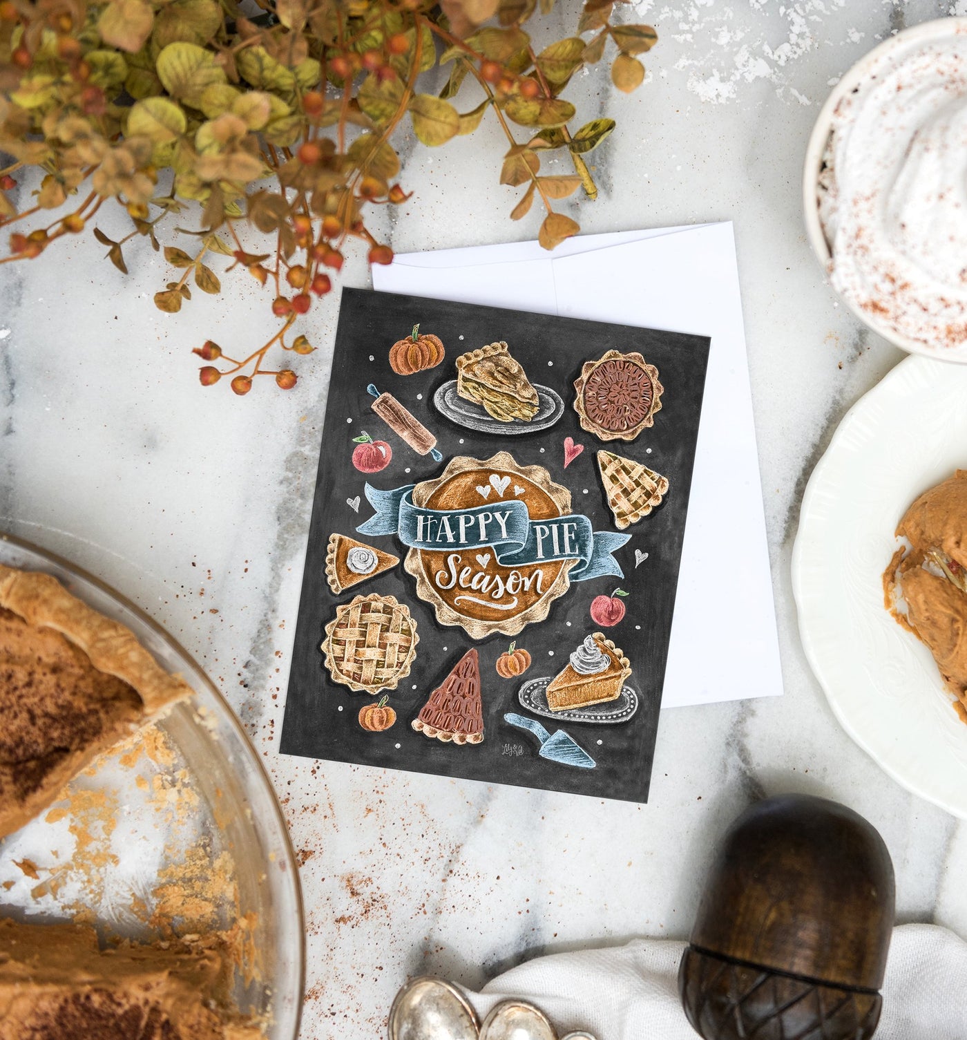 A Happy Pie Season greeting card set amongst thanksgiving day décor which has been placed upon a table with a white table cloth accompanied by none other than a delicious looking pumpkin pie!