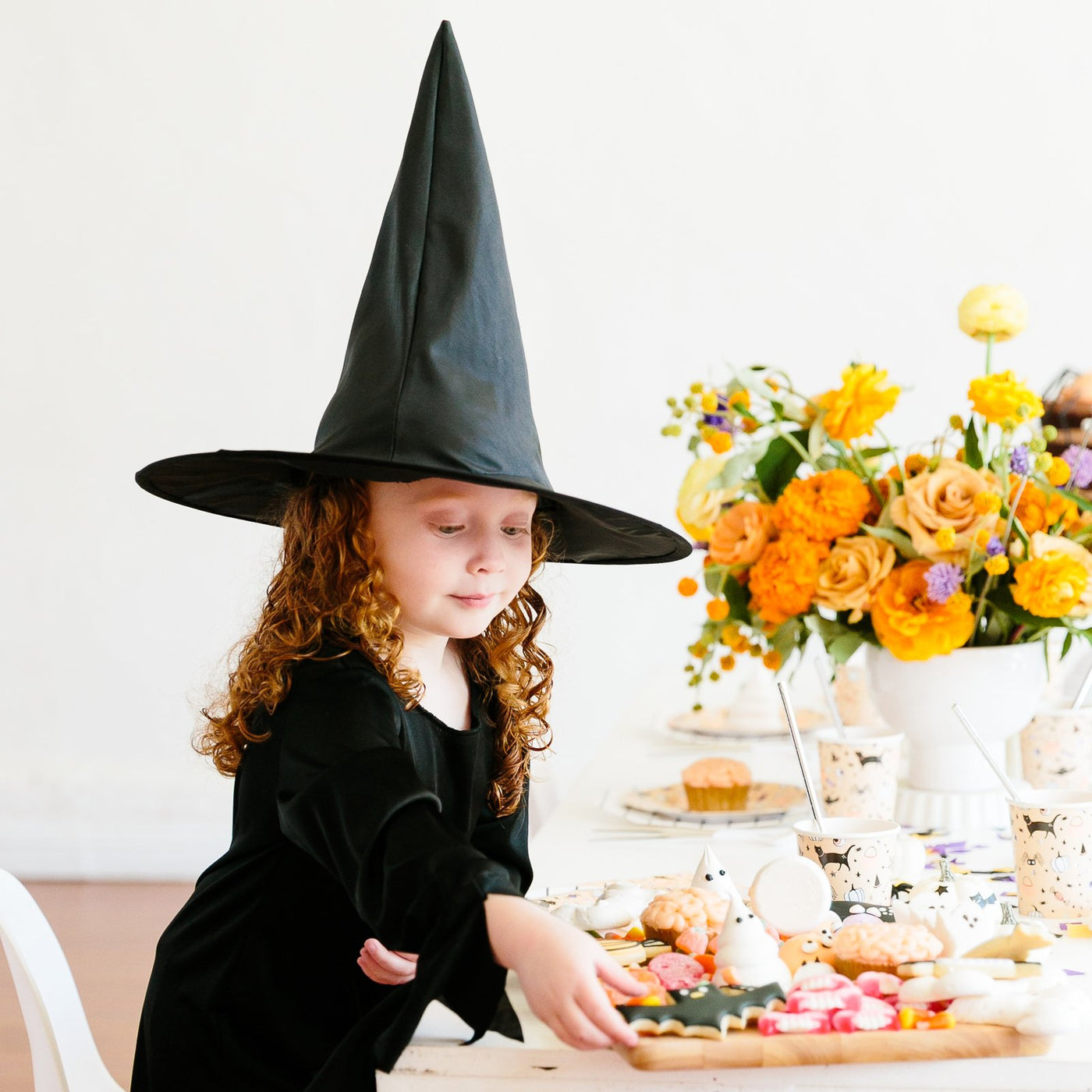 A Little girl dressed in her witch Halloween costume taking a spooky bat Halloween themed cookie from a table with Halloween themed decorations including hocus pocus plates, cups, and large napkins