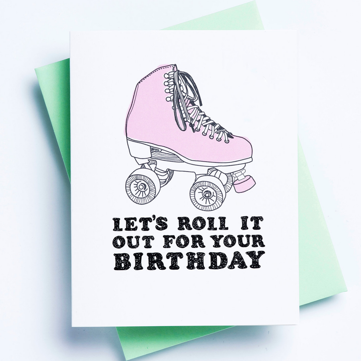 A white birthday card with an image of a roller skate and the words 'let's roll it out for your birthday' displayed over a green envelope and white background