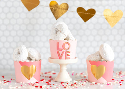 Love baking cups displayed with white donut treats on a table covered in valentines colored sprinkles and a gold heart banner hung in the background